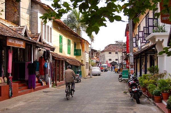 Rows of houses and shops by the side of a road with vehicles parked and a man is seen cycling on the road.